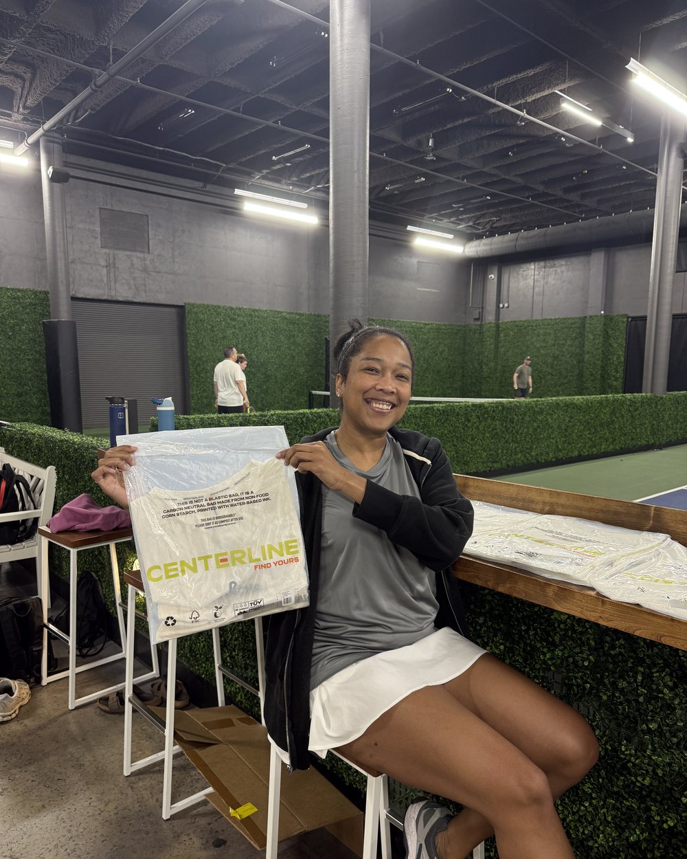 Lauren Bush seated courtside holding a Centerline package, laughing, players on court behind her.