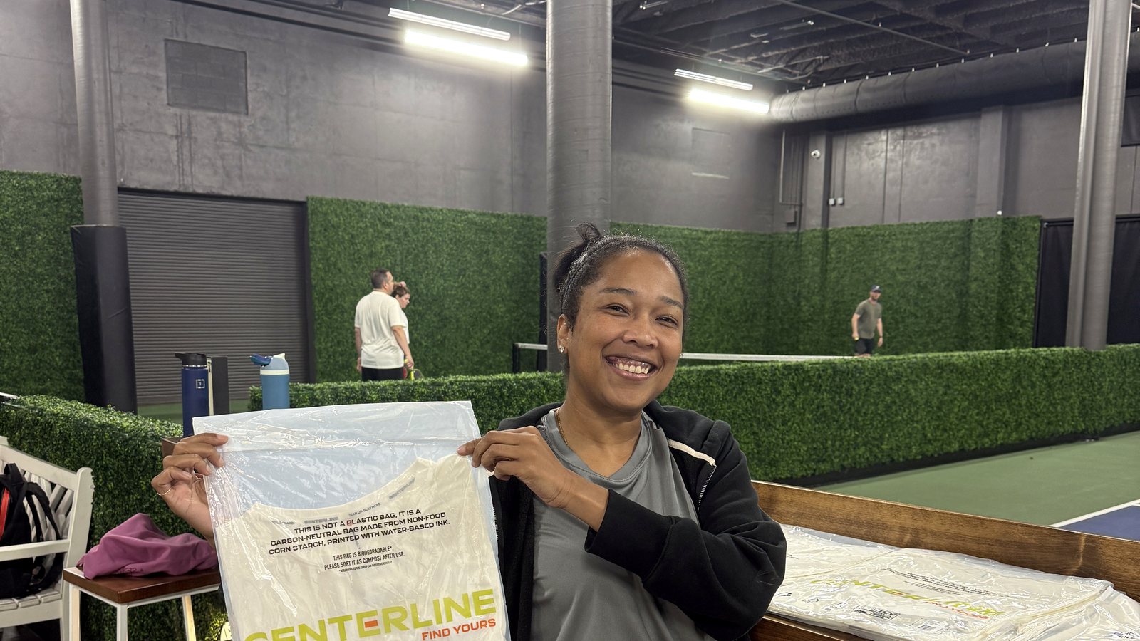 Lauren Bush seated courtside at a Honcho pickleball venue holding up a Centerline apparel package, smiling, with play happening behind her.