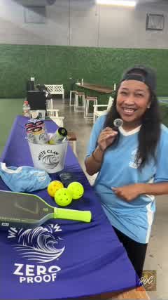 Lauren Bush smiling, holding a handheld microphone at a courtside table stocked with White Claw Zero Proof cans and pickleball gear.